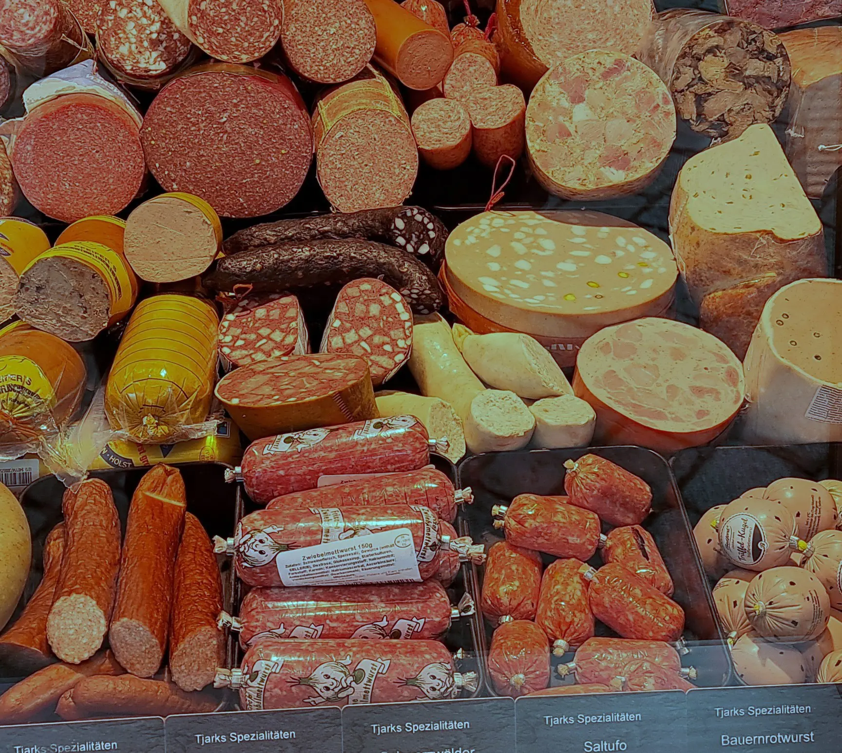 Eine Vitrine, gefüllt mit einer Vielzahl von geschnittenen und ganzen Würsten, Salami und Wurstwaren, die in Reihen angeordnet sind. Die Etiketten unter den Wurstwaren weisen auf die verschiedenen Spezialitäten hin.