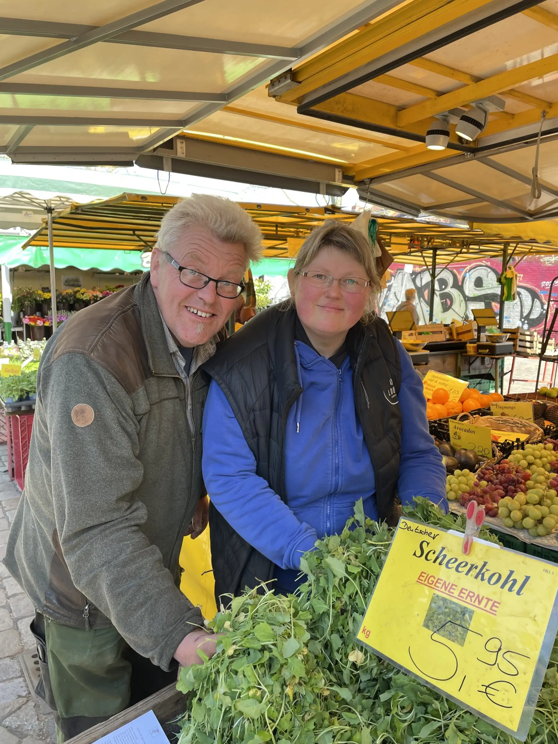 Björn und Inga True stehen hinter einem Marktstand mit frischem Gemüse, darunter ein Haufen Grünzeug mit der Aufschrift Schierkohl. Im Hintergrund sind bunte Produkte und ein gelbes Vordach zu sehen.