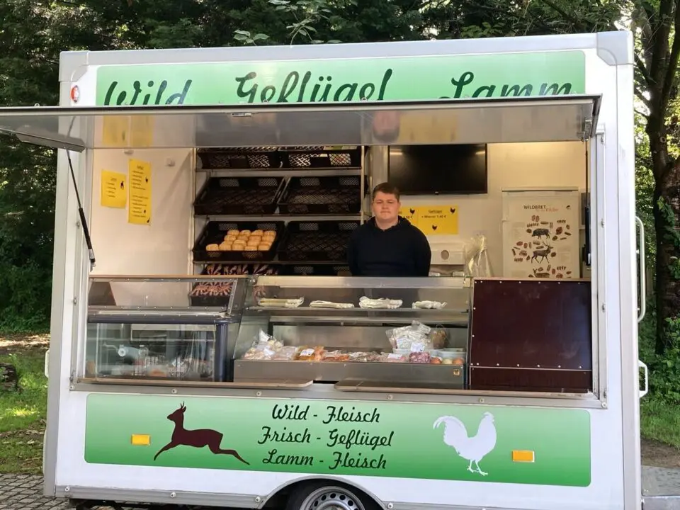 Eine Person steht in einem weißen Lebensmittelwagen, der frische Fleischprodukte verkauft. In den Regalen dahinter sind Brötchen und andere Waren ausgestellt. Der Lkw ist im Freien in der Nähe von Bäumen geparkt, mit einem grünen Schild, auf dem ein Hirsch und ein Hahn abgebildet sind.