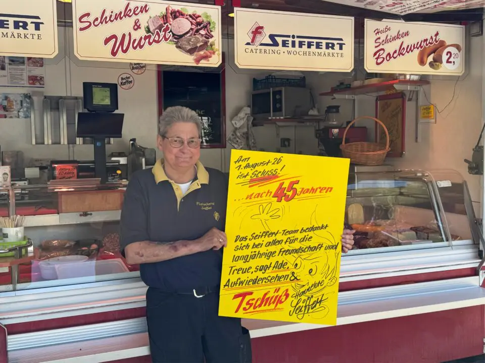 Hannelore Seiffert steht vor einer Feinkosttheke und hält ein gelbes Abschiedsschild in der Hand. Auf dem Schild kündigt sie ihren Ruhestand nach 45 Jahren bei Seiffert Schinken & Wurst an und bedankt sich bei den Kunden für ihre Treue.