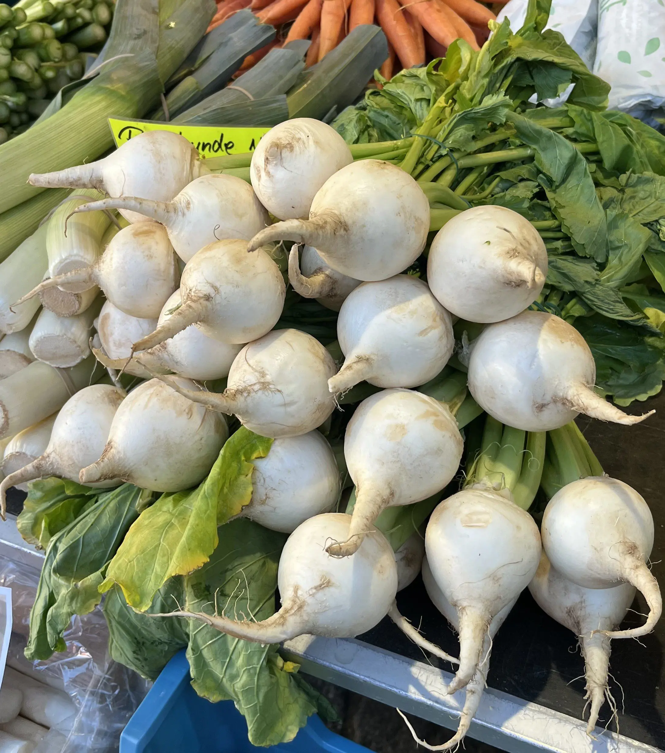 Ein Haufen frischer weißer Rüben mit grünen Blattspitzen auf einem Markt, umgeben von anderem Gemüse wie Karotten und Mais im Hintergrund.