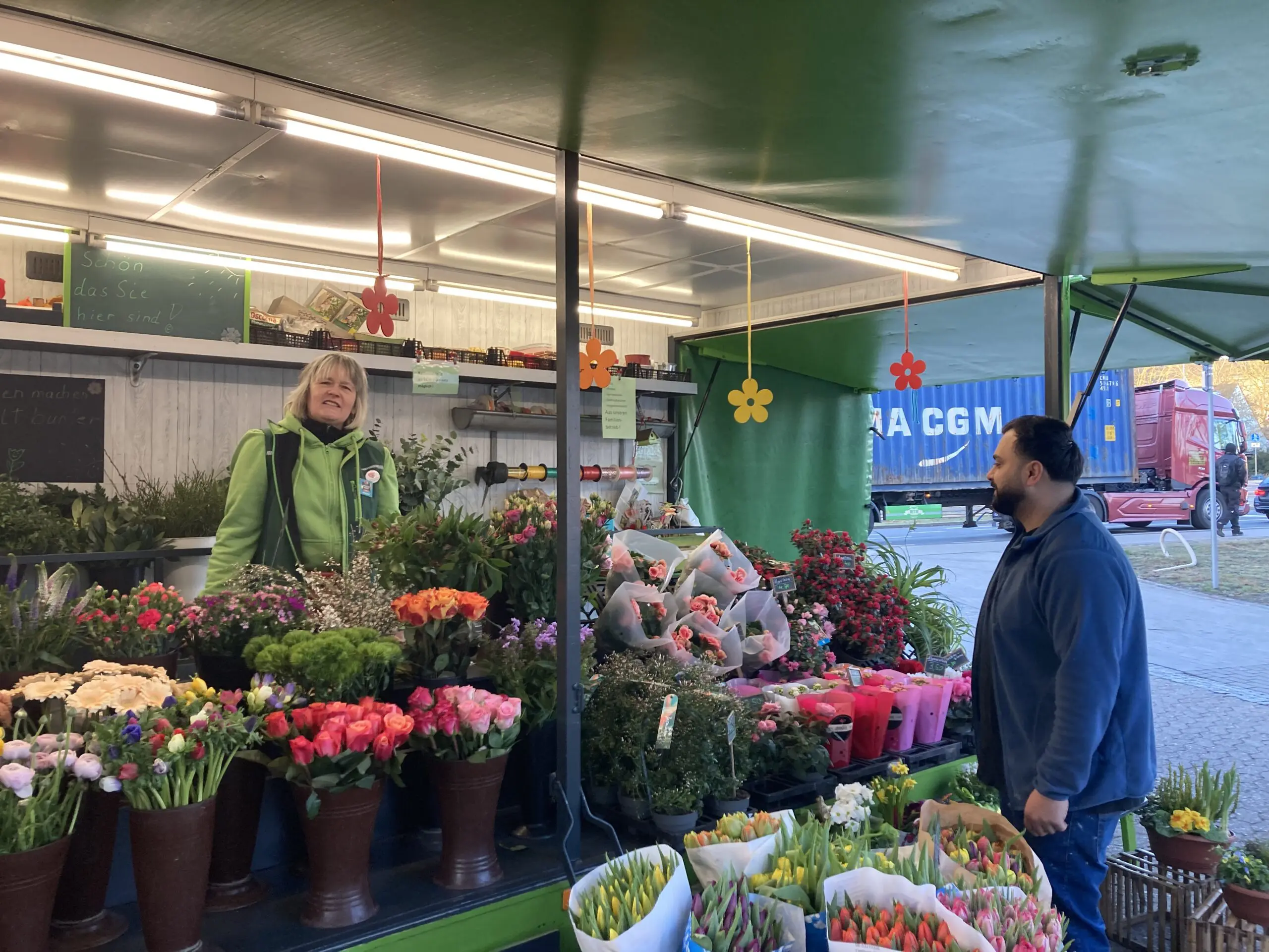 Imke Schulenberg steht hinter dem Tresen eines Blumenladens mit bunten Sträußen und Pflanzen und unterhält sich mit einem Mann, der an einem sonnigen Tag im Freien vor dem Laden steht.