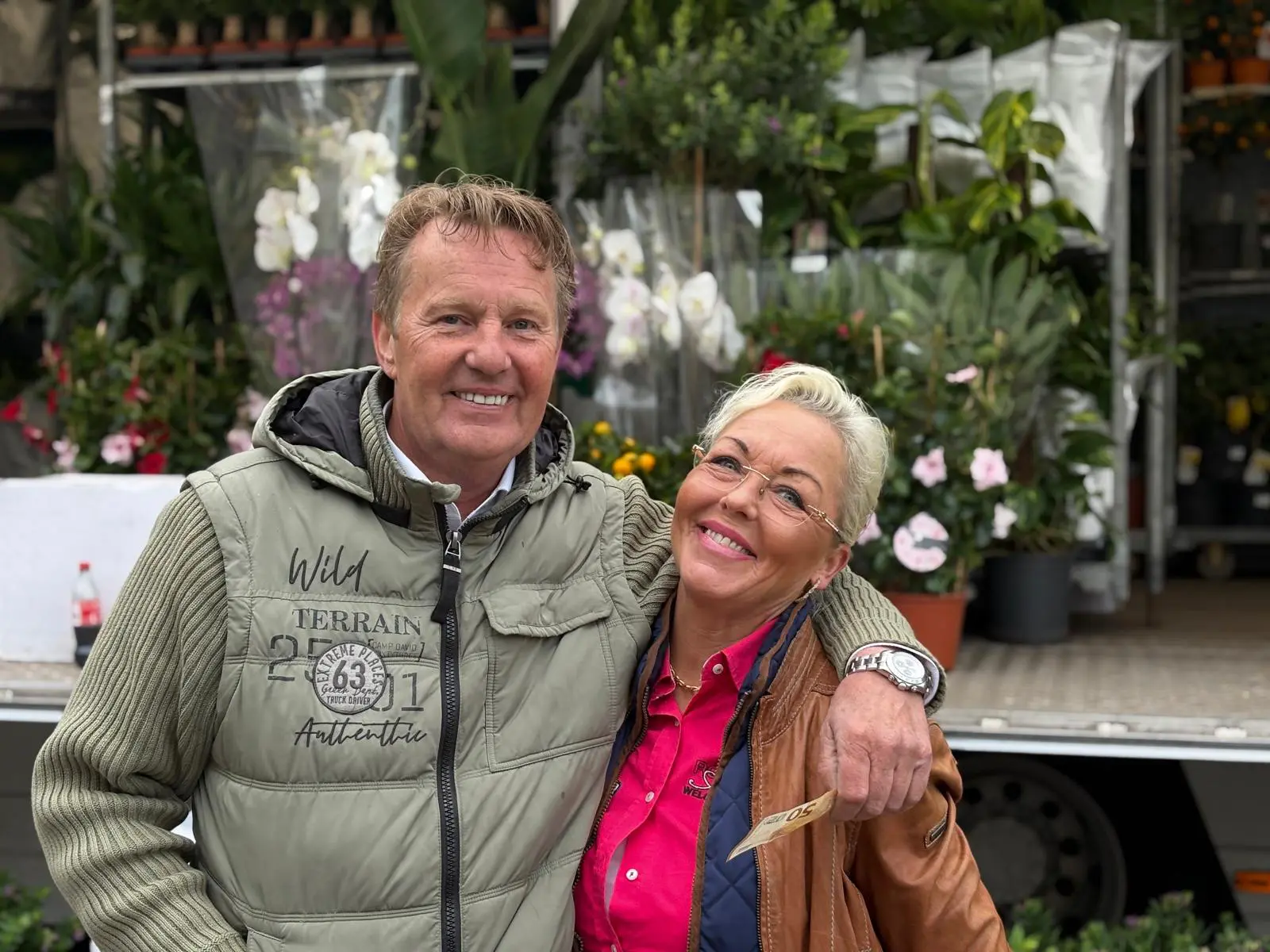 Ein lächelnder Mann und eine lächelnde Frau stehen dicht beieinander vor einem Blumenstand, der mit verschiedenen Pflanzen und Blumen gefüllt ist. Der Mann trägt eine grüne Jacke, und die Frau hält eine Banknote, während sie eine Brille und eine braune Jacke trägt. Michel und Renata Saarloos