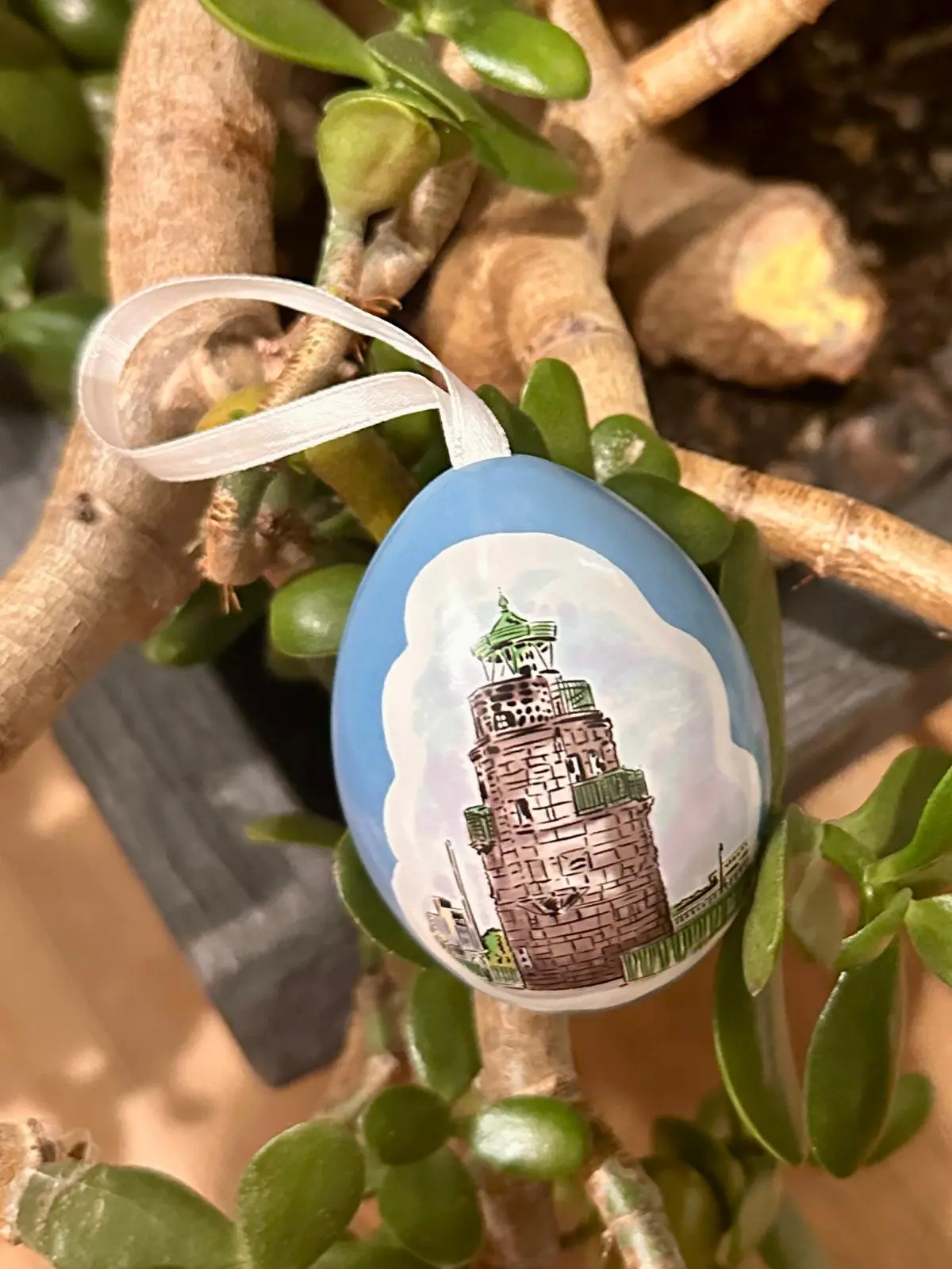 Ein blaues eiförmiges Ornament mit einem weißen Band hängt an einer Jadepflanze. Das Ornament zeigt die Illustration eines steinernen Leuchtturms mit grünem Dach vor einem wolkenverhangenen Himmel.