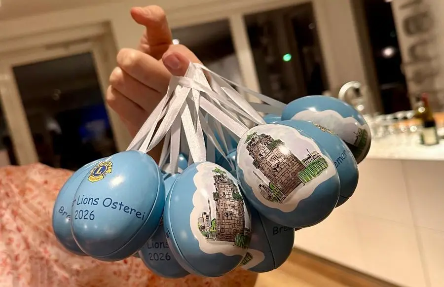 Eine Person hält mehrere blaue runde Ornamente mit weißen Bändern, auf denen Abbildungen von Gebäuden und der Text Lions Osterei 2026 Braunschweig zu sehen sind. Die Ornamente sind in einem gut beleuchteten Innenraum ausgestellt.