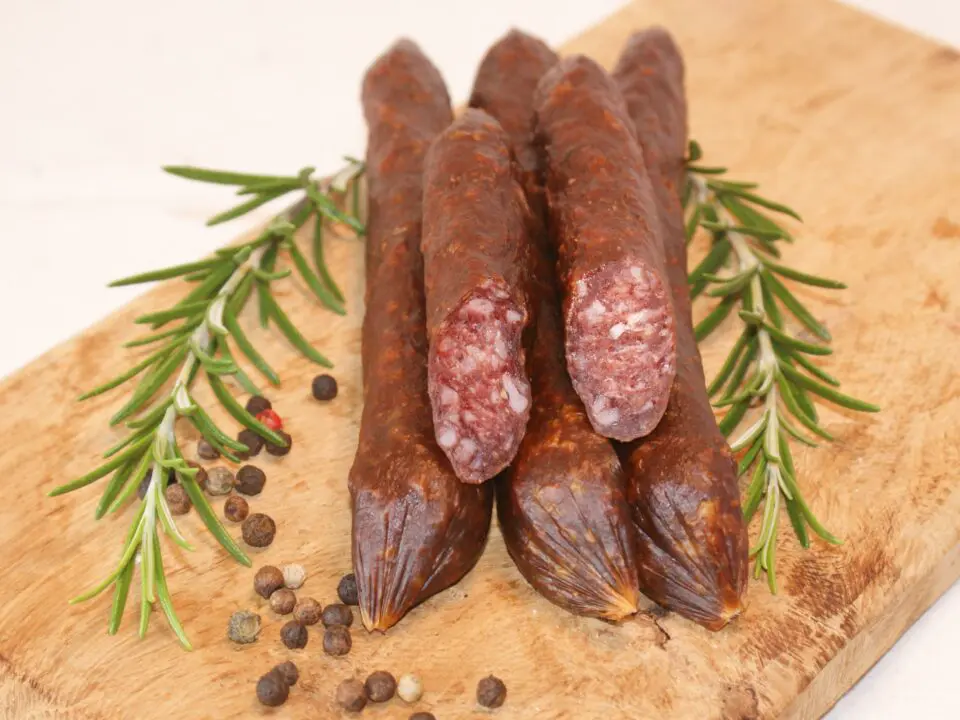 Drei getrocknete Wildwürste liegen auf einem Holzbrett, flankiert von frischen Rosmarinzweigen und gestreuten Pfefferkörnern. Eine Wurst ist aufgeschnitten, um die innere Struktur zu zeigen - eine perfekte Ergänzung für jeden Aufstrich im Bremer Marktkalender.