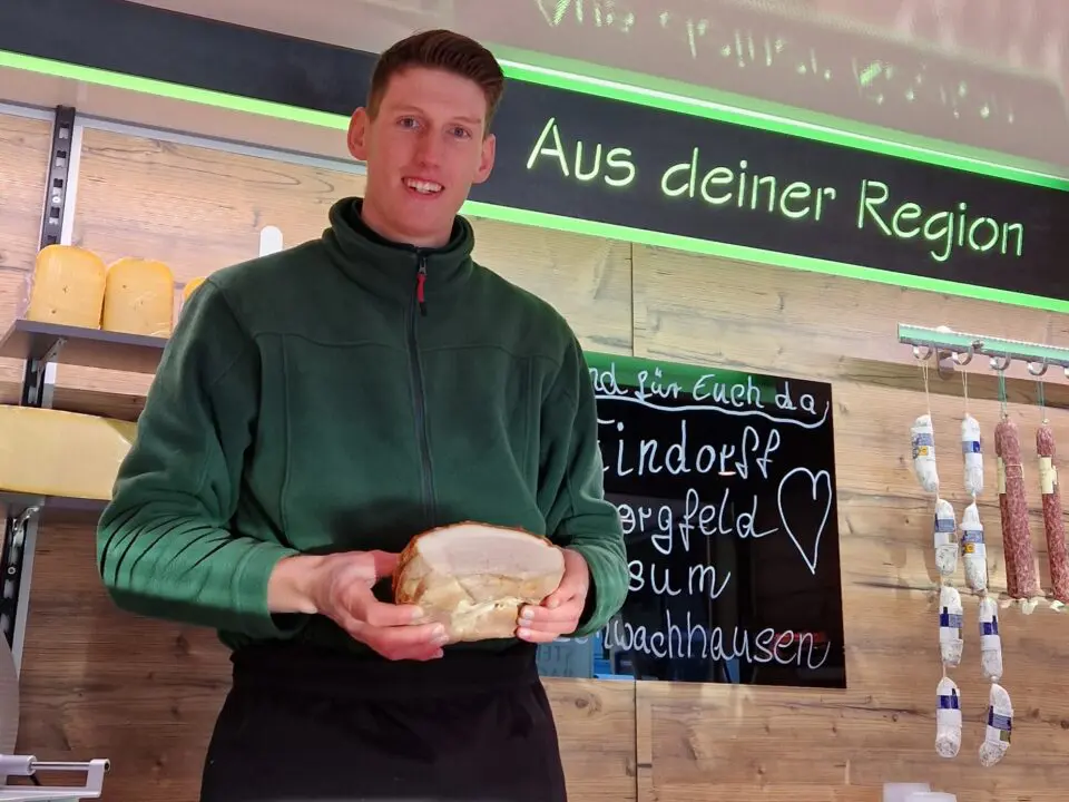 Ein lächelnder Mann Tjark Beckmann in einem grünen Fleece steht hinter einer Feinkosttheke und hält ein großes Stück Fleisch in der Hand. Hinter ihm stehen Regale mit Käse und Wurstwaren, und auf einer Kreidetafel steht "Aus deiner Region".