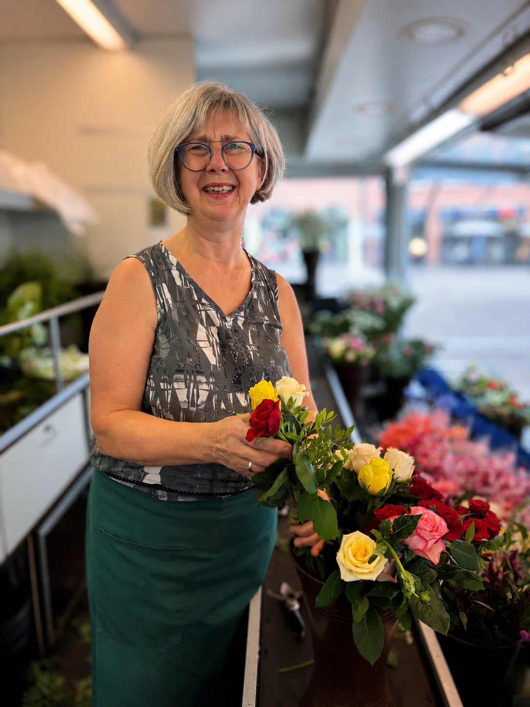 Eine lächelnde Frau mit grauen Haaren (Silvia Maring) arrangiert in einem Blumenladen einen Strauß bunter Rosen. Sie trägt eine Brille, ein gemustertes, ärmelloses Oberteil und eine grüne Schürze. Im Hintergrund sind leuchtende Blumen und Grünpflanzen zu sehen.