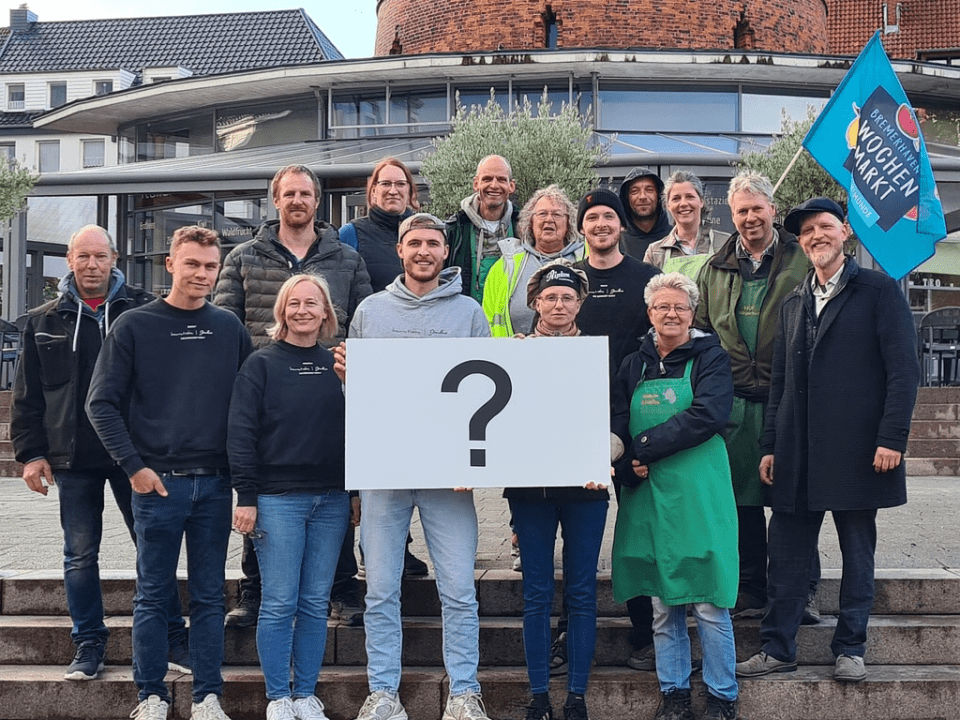 Eine Gruppe von Menschen steht auf einer Freitreppe, lächelt und posiert zusammen. Zwei Personen vor ihnen halten ein großes weißes Schild mit einem Fragezeichen. Im Hintergrund sind eine blaue Flagge und ein runder Backsteinturm zu sehen.