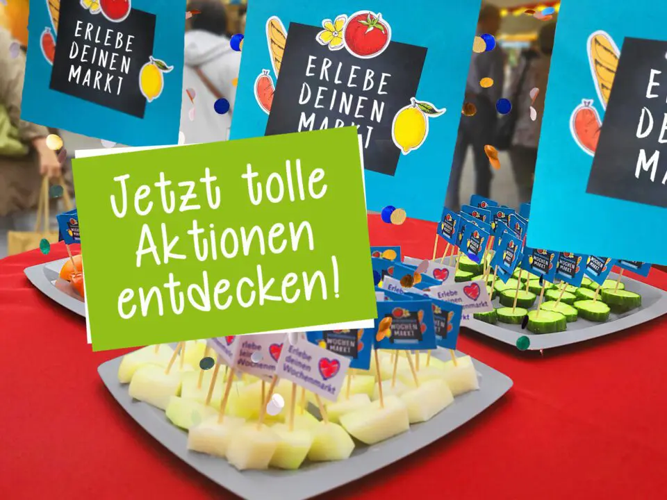 Eine Auslage mit Obst- und Gemüseproben auf Tabletts mit kleinen Schildern, die auf einem roten Tisch auf einem Markt aufgestellt sind. Auf den Schildern steht "Erlebe deinen Markt" und "Jetzt tolle Aktionen entdecken!" auf Deutsch. Im unscharfen Hintergrund sind Menschen zu sehen.