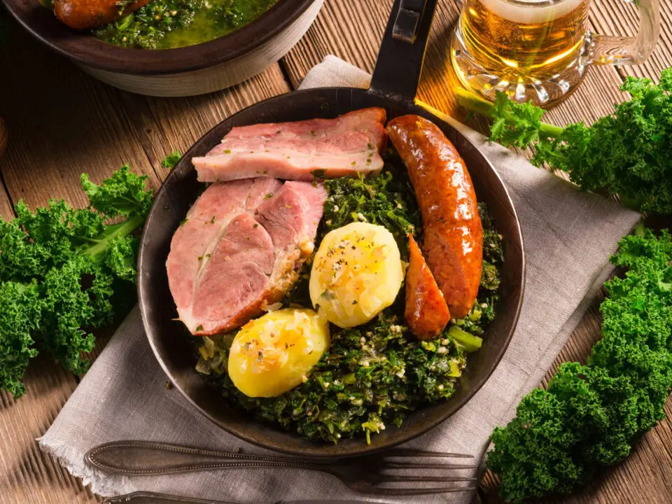 Ein rustikaler Teller mit gekochten Kartoffeln, Schinken, Wurst und gekochtem Grünkohl auf einem Holztisch, garniert mit frischem Grünkohl. Im Hintergrund stehen ein Krug Bier und ein weiterer Teller mit Wurst und Grünkohl.