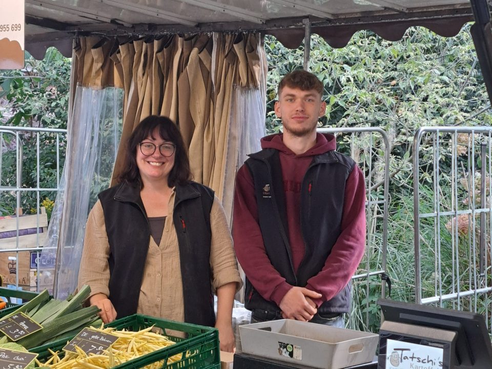 Zwei Marktverkäufer, eine Frau und ein Mann, stehen lächelnd hinter einem Verkaufsstand mit frischem Gemüse wie Bohnen, Lauch und grünen Zwiebeln. Auf der Theke stehen eine Geldkassette und ein Schild mit der Aufschrift "Tatschis Kartoffeln".