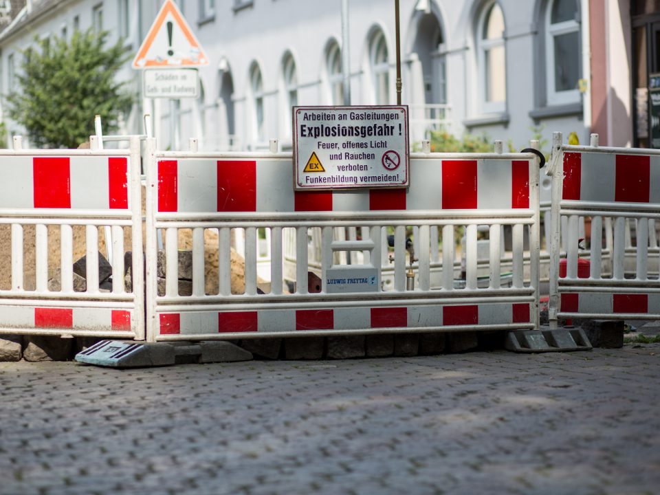 Eine Baustelle auf einer Kopfsteinpflasterstraße ist von rot-weißen Absperrungen umgeben. Ein Warnschild weist auf die Explosionsgefahr bei Arbeiten an einer Gasleitung hin. In der Nähe befinden sich Wohngebäude und ein Straßenschild.