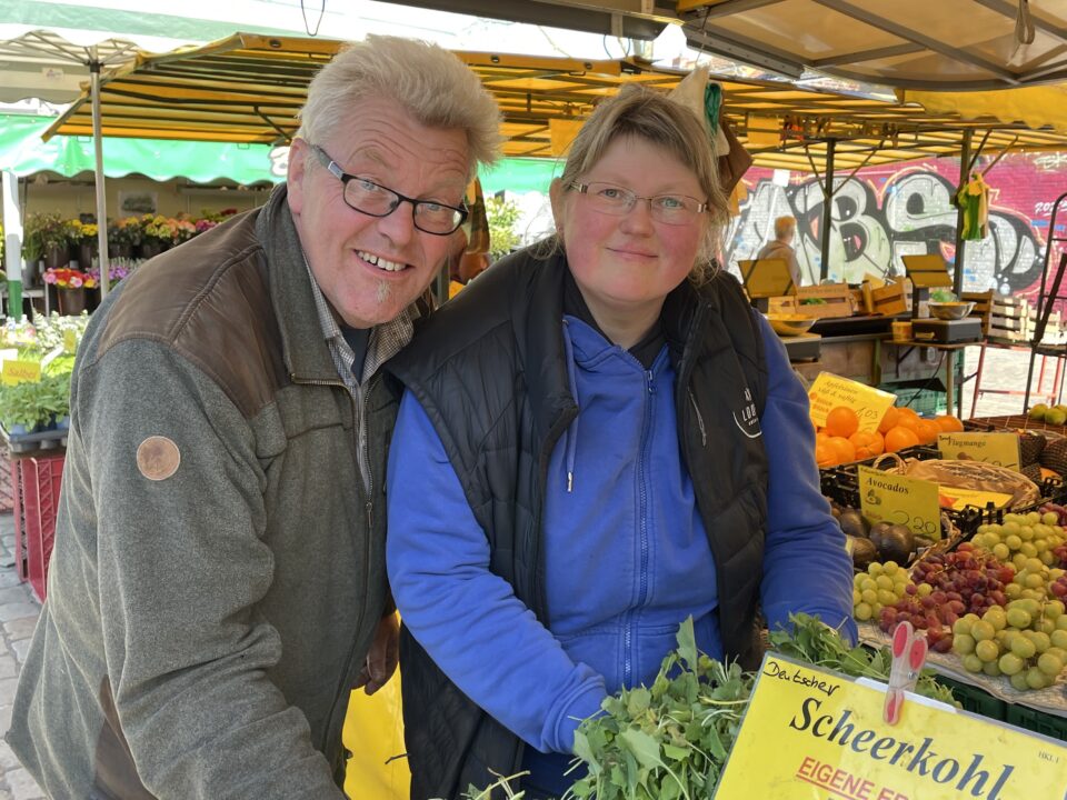 Ein lächelnder Mann und eine lächelnde Frau stehen an einem Marktstand und verkaufen Gemüse, Weintrauben und Orangen. Gelbe Preisschilder in deutscher Sprache sind zu sehen. Der Markt findet im Freien statt, mit bunten Zelten und Graffiti im Hintergrund.