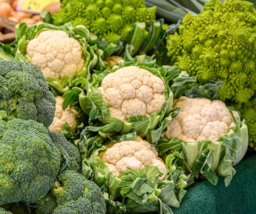Eine Auslage mit frischem grünen Brokkoli, weißem Blumenkohl und leuchtend grünem Romanesco auf einem Markt, wobei das Gemüse dicht nebeneinander auf einer grünen Fläche angeordnet ist.