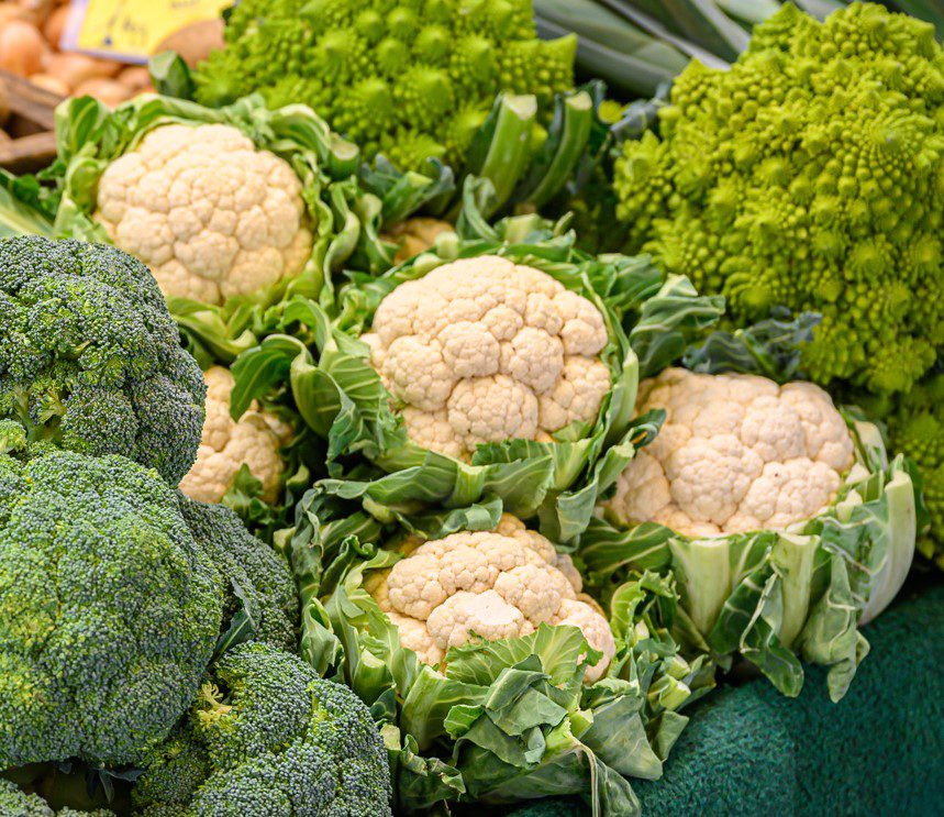 Eine Auslage mit frischem grünen Brokkoli, weißem Blumenkohl und leuchtend grünem Romanesco auf einem Markt, wobei das Gemüse dicht nebeneinander auf einer grünen Fläche angeordnet ist.