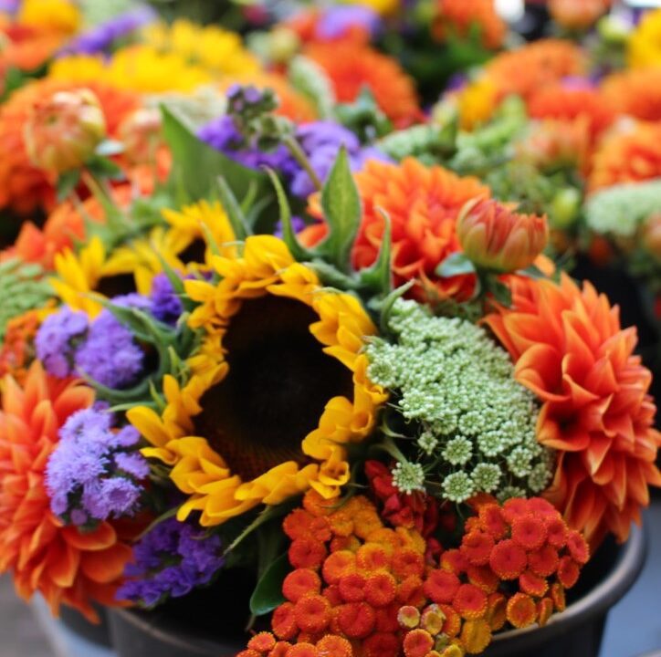 Ein farbenfrohes Blumenbouquet aus Sonnenblumen, orangefarbenen Dahlien sowie kleinen violetten, weißen und roten Blüten, arrangiert in einem schwarzen Eimer, während im unscharfen Hintergrund weitere ähnliche Blumenarrangements zu sehen sind.
