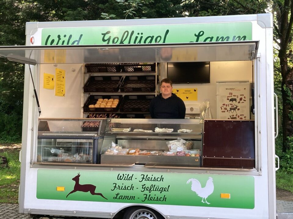 Eine Person steht in einem weißen Lebensmittelwagen, der frische Fleischprodukte verkauft. In den Regalen dahinter sind Brötchen und andere Waren ausgestellt. Der Lkw ist im Freien in der Nähe von Bäumen geparkt, mit einem grünen Schild, auf dem ein Hirsch und ein Hahn abgebildet sind.