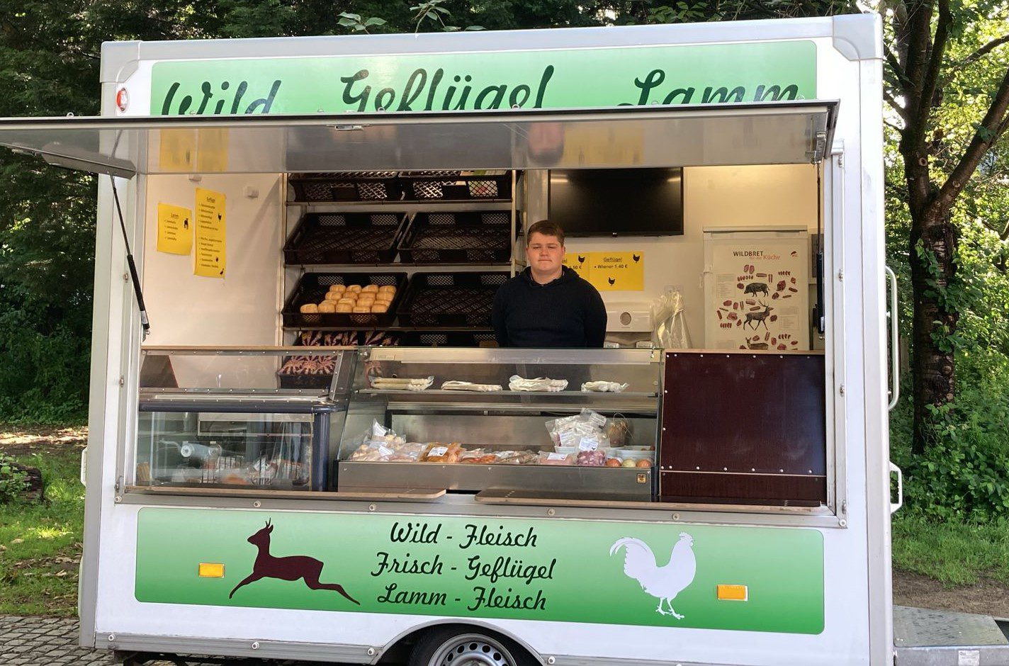 Ein Mann steht in einem grün-weißen Imbisswagen und verkauft frisches Fleisch und Brötchen. In der Vitrine sind verschiedene Fleischprodukte ausgestellt, und auf Schildern sind Optionen wie Wild, Geflügel und Lamm aufgeführt. Im Hintergrund sind Bäume zu sehen.