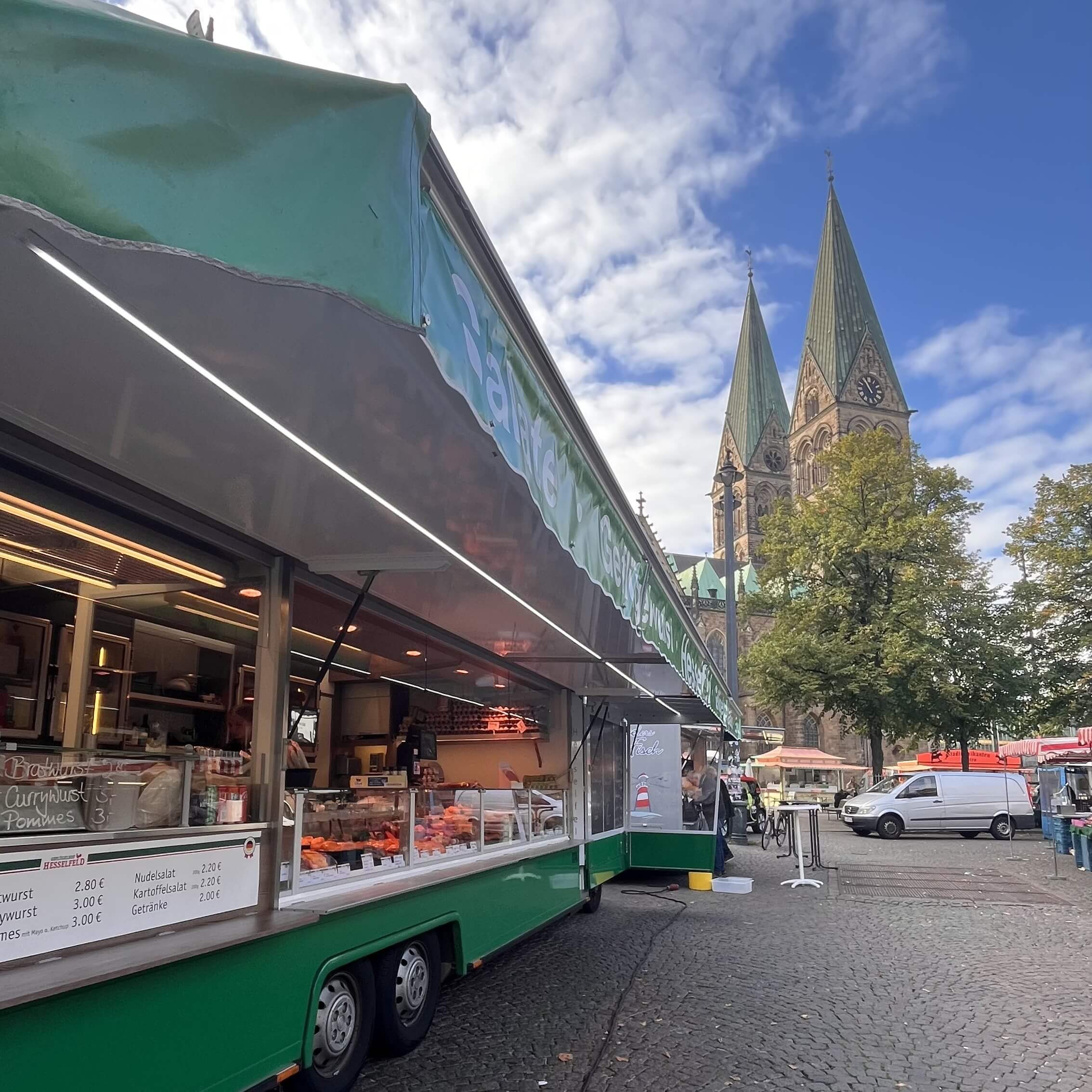 Ein grüner Wochenmarktwagen verkauft Geflügelprodukte auf dem Wochenmarkt Domshof unter freiem Himmel. Im Hintergrund erhebt sich unter einem teilweise bewölkten Himmel eine historische Kirche mit zwei hohen Türmen. Die Menschen besuchen die nahegelegenen Marktstände auf dem Kopfsteinpflasterplatz.