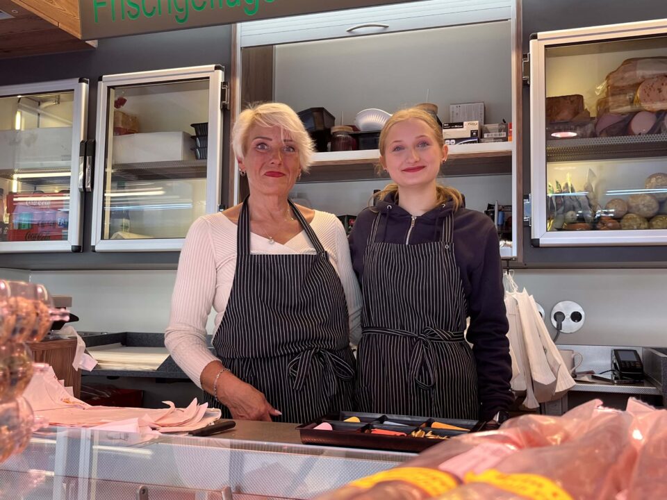 Zwei Frauen in gestreiften Schürzen stehen hinter einer Feinkosttheke mit Aufschnitt und Käsescheiben und lächeln in die Kamera. Im Hintergrund sind Regale mit Aufschnitt und Feinkostprodukten zu sehen.