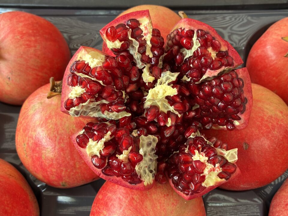 Ein aufgeschnittenes Granatapfel, aus dem die roten Kerne hervorschauen, umgeben von mehreren ganzen Granatäpfeln in einer Plastikschale.