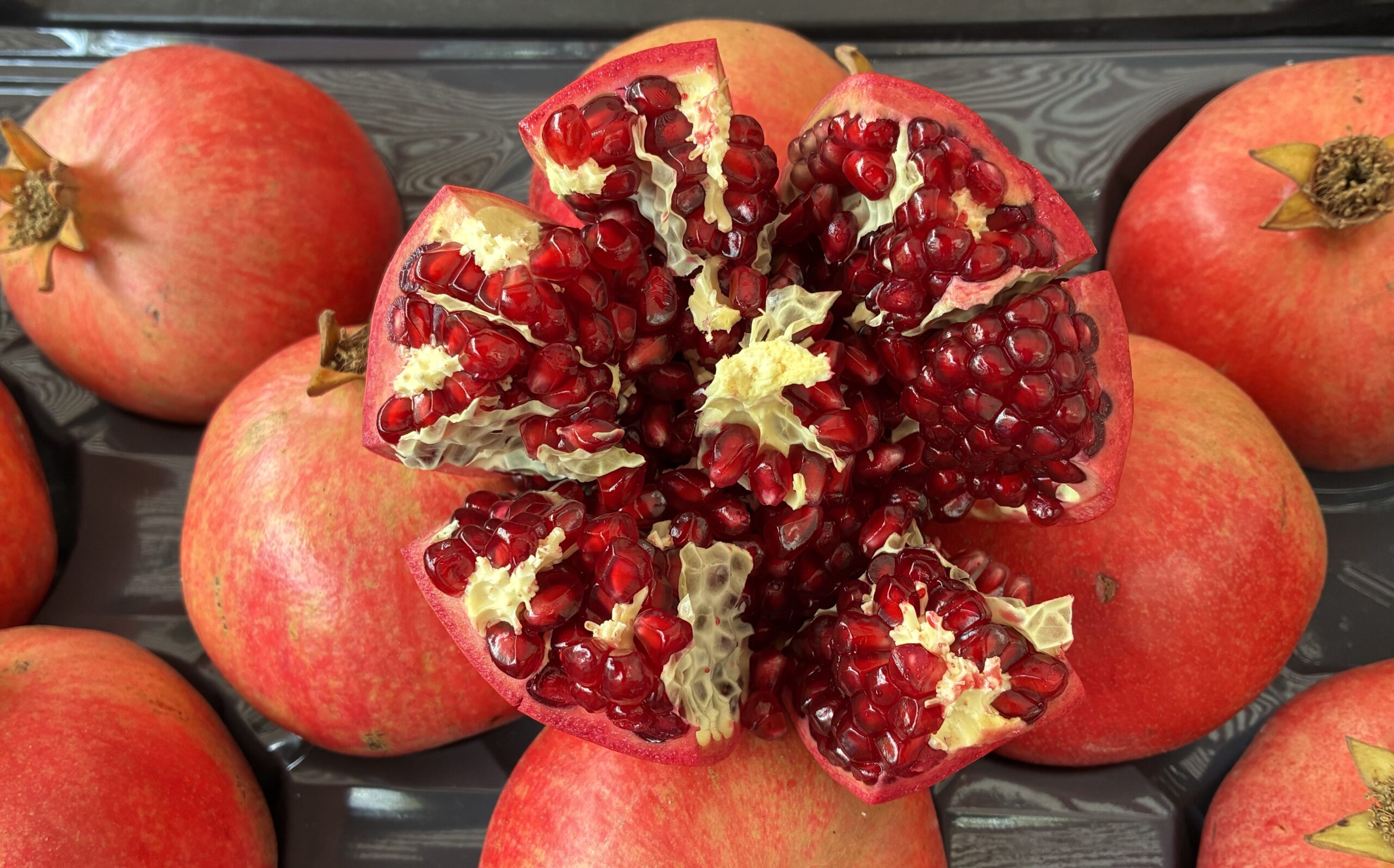 Ein aufgeschnittenes Granatapfel, aus dem die roten Kerne hervorschauen, umgeben von mehreren ganzen Granatäpfeln in einer Plastikschale.