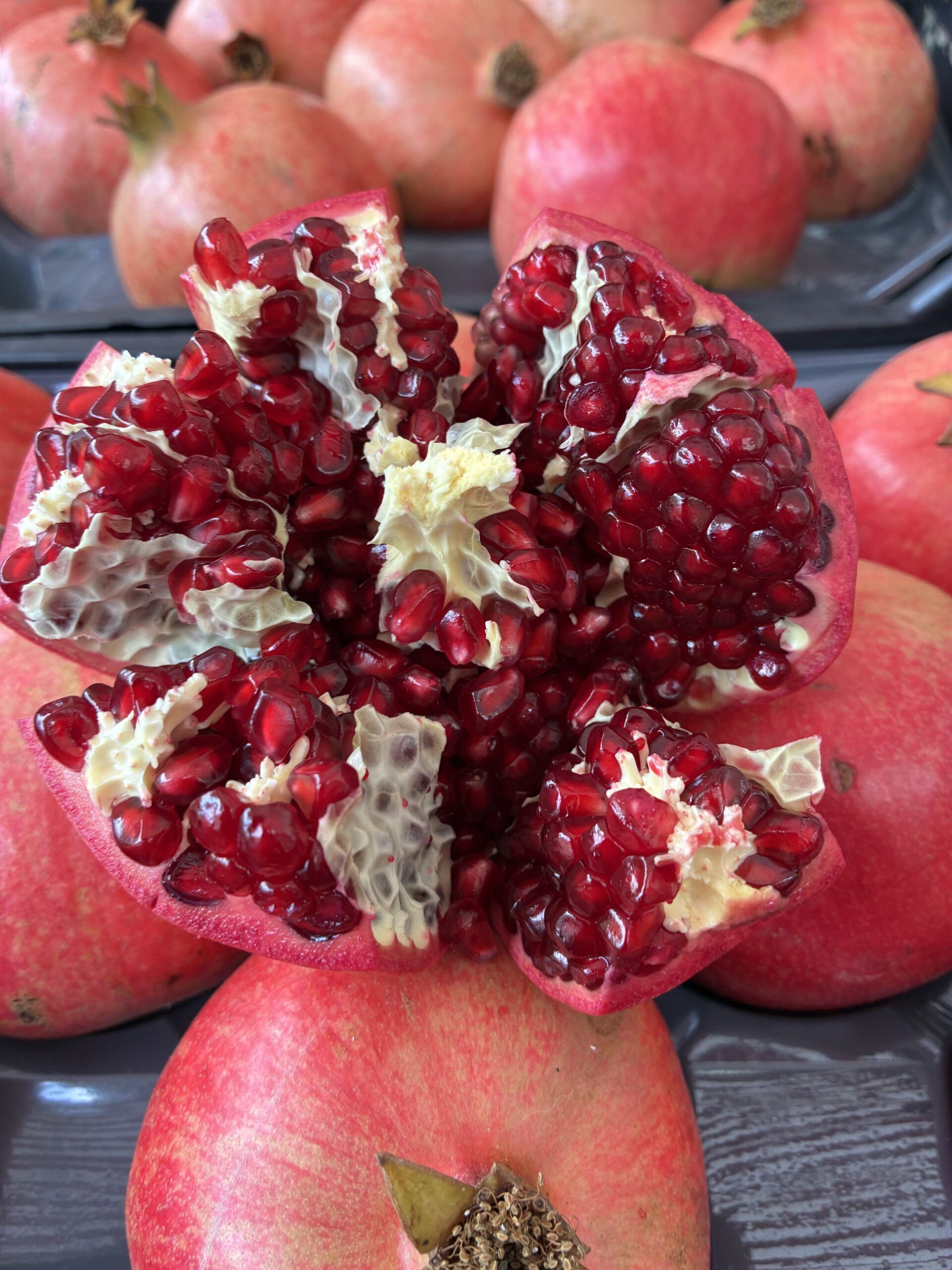 Ein aufgeschnittenes Granatapfel, aus dem die roten Kerne hervorschauen, umgeben von ganzen Granatäpfeln in einer Plastikschale.