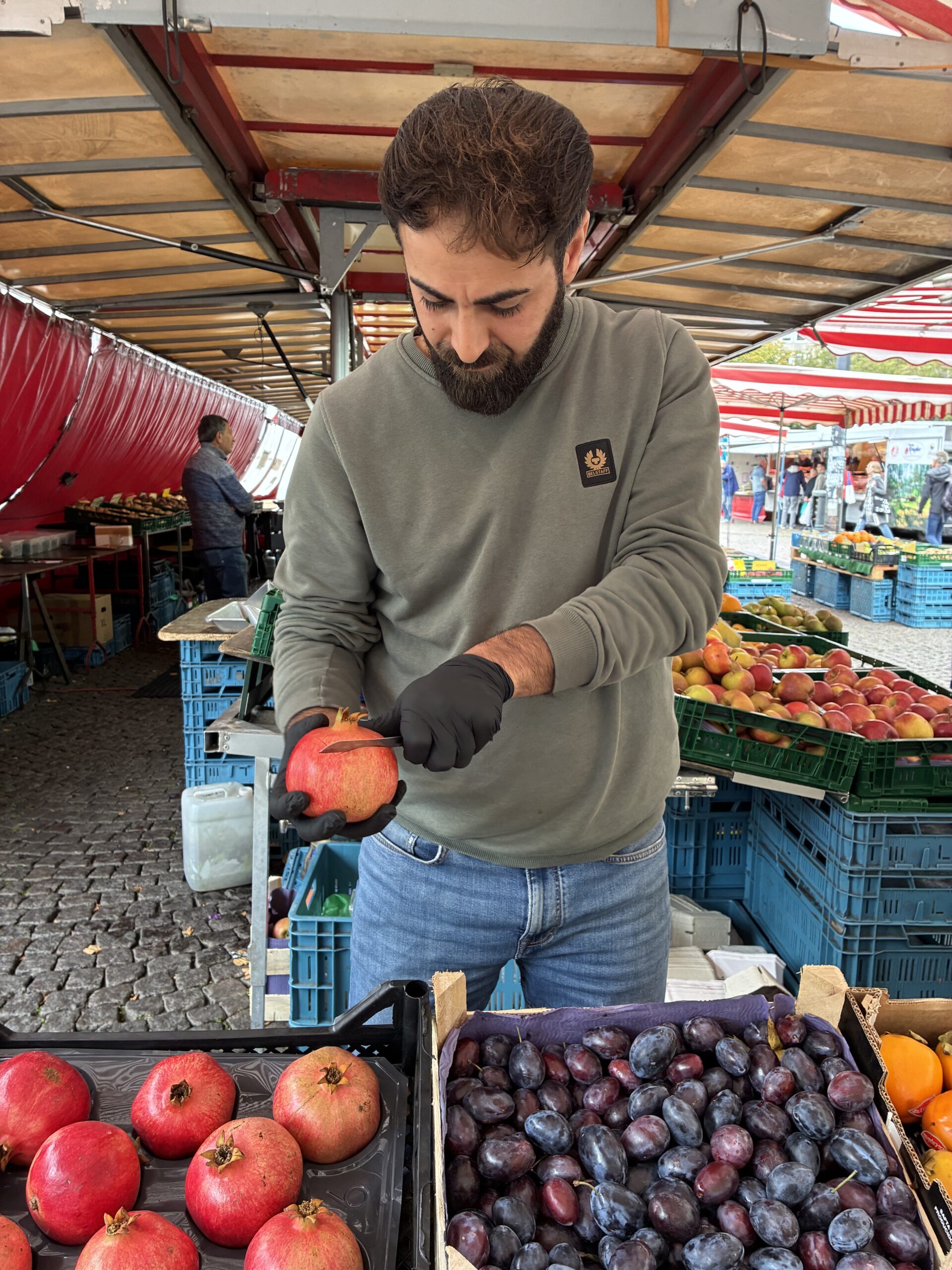 Ein Mann in einem grünen Sweatshirt und schwarzen Handschuhen schneidet auf einem Markt im Freien mit einem Messer einen Granatapfel auf.