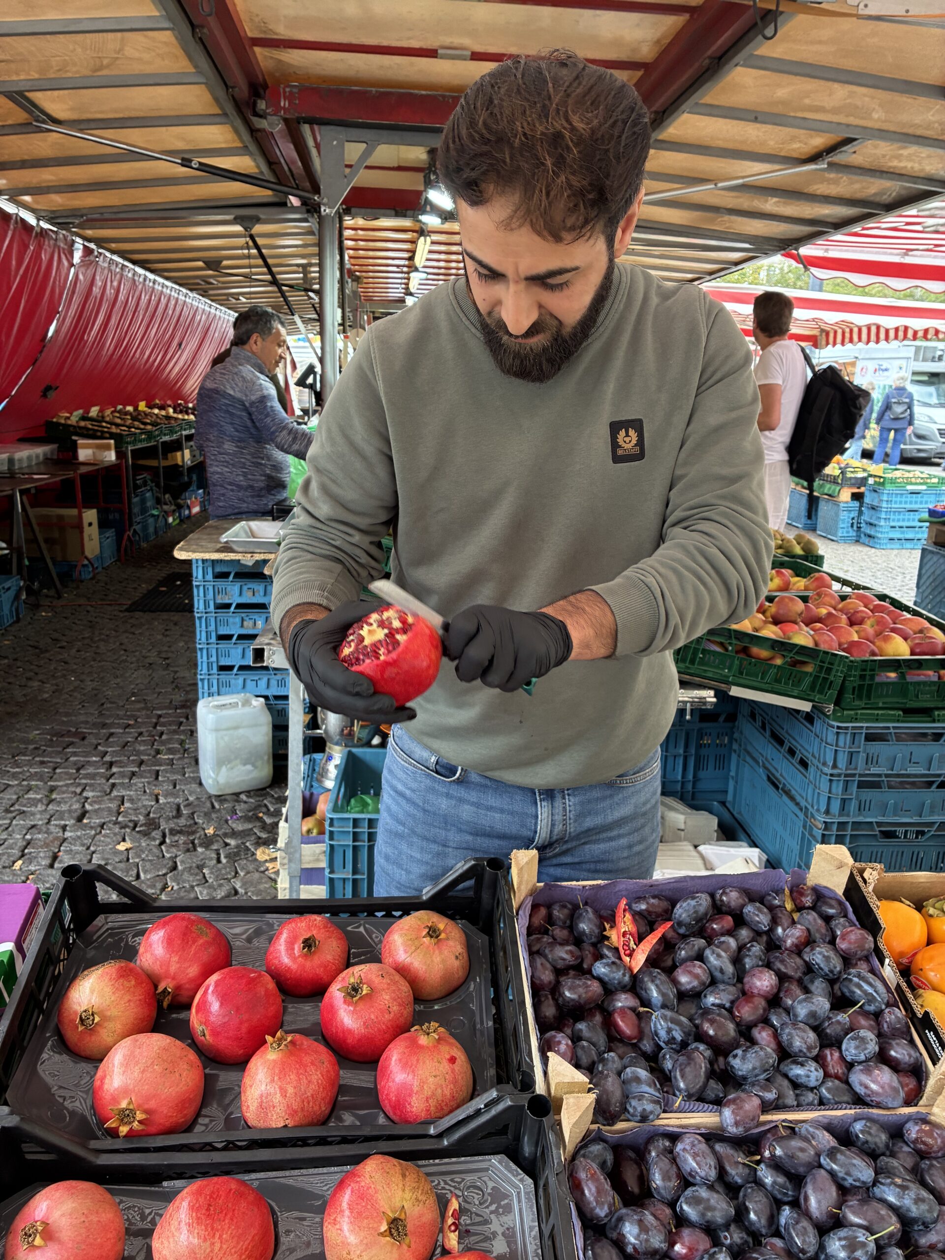 Ein bärtiger Marktverkäufer mit schwarzen Handschuhen schneidet an einem Obststand, der mit Granatäpfeln und Pflaumen gefüllt ist, einen Granatapfel auf.