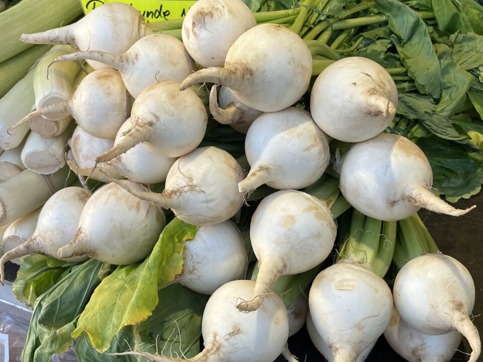 Ein Haufen frischer weißer Rüben mit grünen Blattspitzen auf einem Markt, umgeben von anderem Gemüse wie Karotten und Mais im Hintergrund.