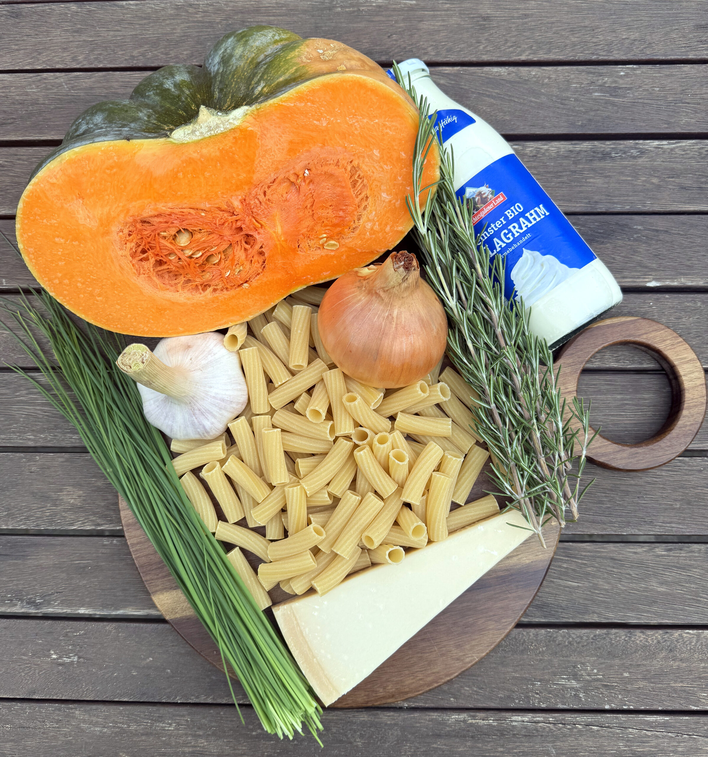 Ein Holzbrett mit Rigatoni, einem Stück Käse, Schnittlauch, Knoblauch, Zwiebeln, Rosmarin, einem halbierten Kürbis und einer Flasche Sahne auf einem Holztisch.