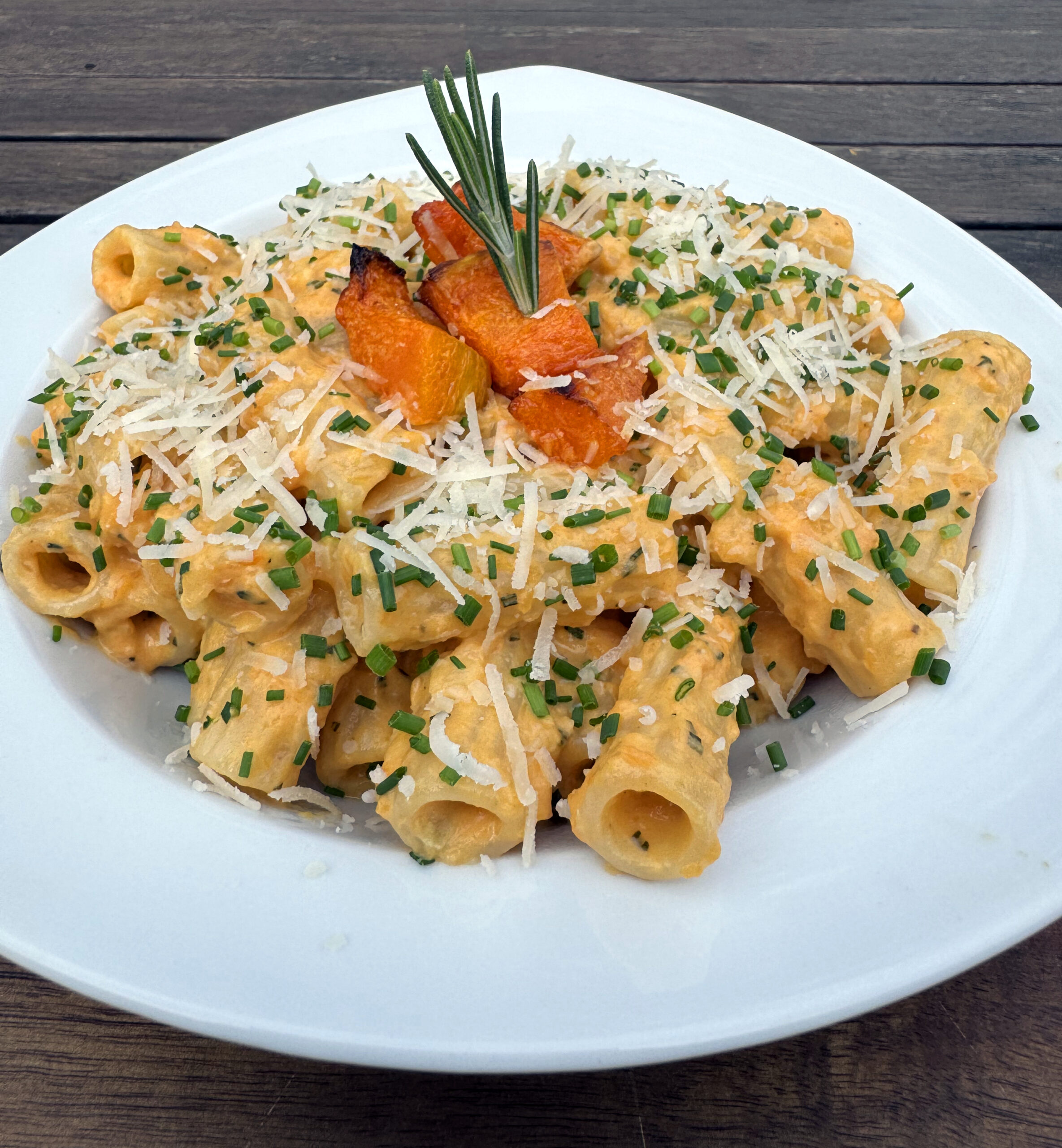 Ein weißer Teller mit Rigatoni in einer cremigen Orangensoße, garniert mit geriebenem Käse, gehacktem Schnittlauch, gerösteten Kürbisstücken und einem Rosmarinzweig, steht auf einem Holztisch.
