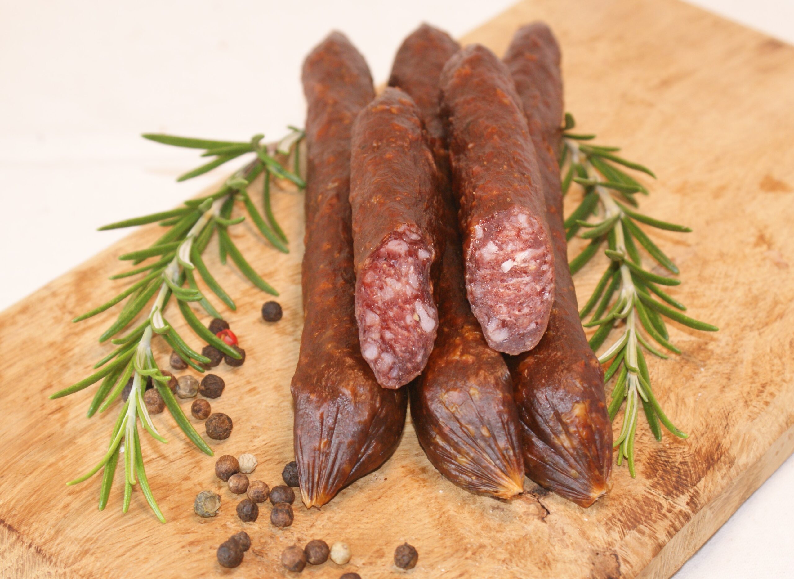 Drei getrocknete Wildwürste liegen auf einem Holzbrett, flankiert von frischen Rosmarinzweigen und gestreuten Pfefferkörnern. Eine Wurst ist aufgeschnitten, um die innere Struktur zu zeigen - eine perfekte Ergänzung für jeden Aufstrich im Bremer Marktkalender.
