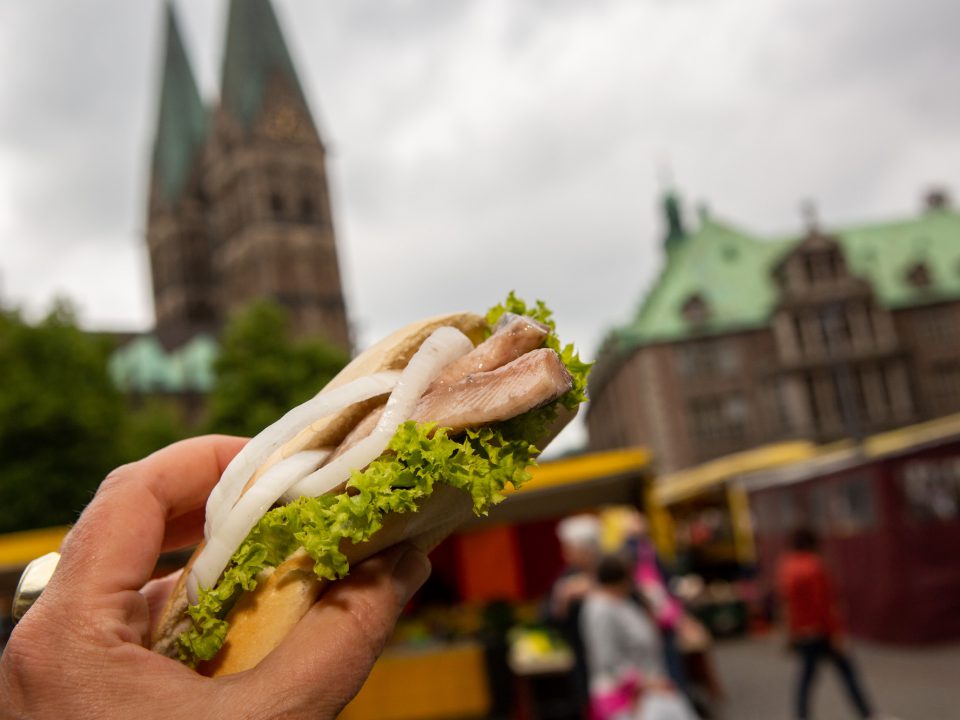 Matjesbrötchen wird in der Hand gehalten; im Hintergrund ist der Bremer Dom zu sehen.