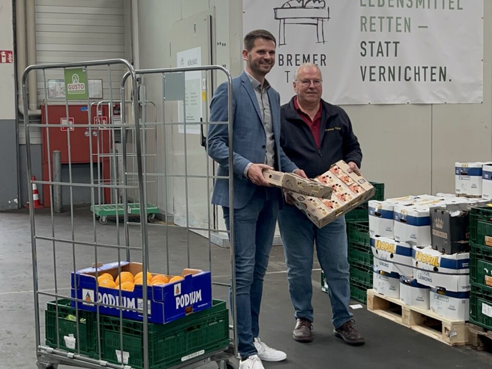 Zwei Männer (Lars Jansen vom Großmarkt und Uwe Schneider von der Tafel) stehen in einer Lagerhalle neben Lebensmittelkisten und einem Schild mit der Aufschrift „Lebensmittel retten – statt vernichten“. Sie lächeln und halten verpackte Waren in den Händen, umgeben von Kisten mit Lebensmitteln.