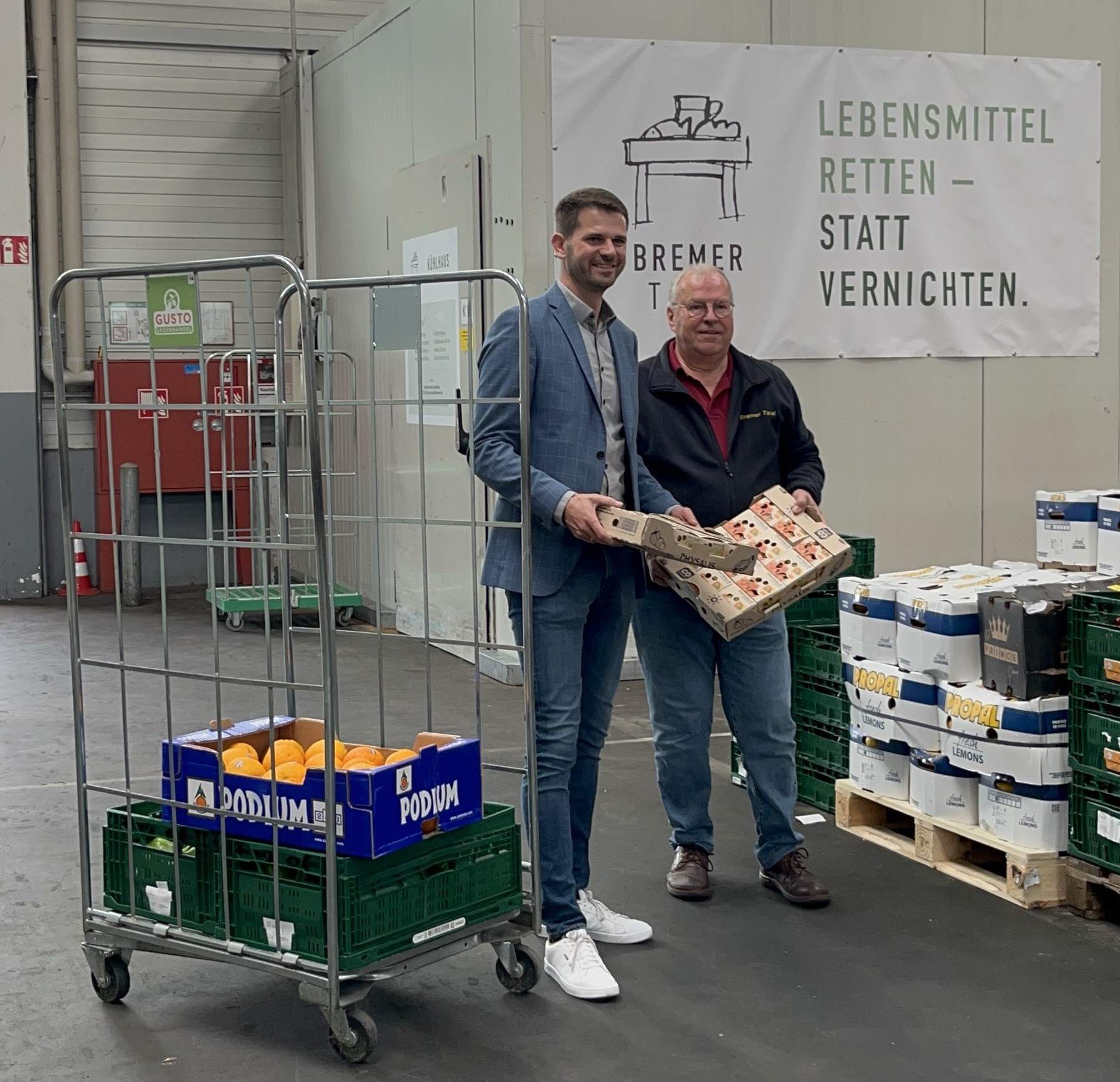 Zwei Männer (Lars Jansen vom Großmarkt und Uwe Schneider von der Tafel) stehen in einer Lagerhalle neben Lebensmittelkisten und einem Schild mit der Aufschrift „Lebensmittel retten – statt vernichten“. Sie lächeln und halten verpackte Waren in den Händen, umgeben von Kisten mit Lebensmitteln.
