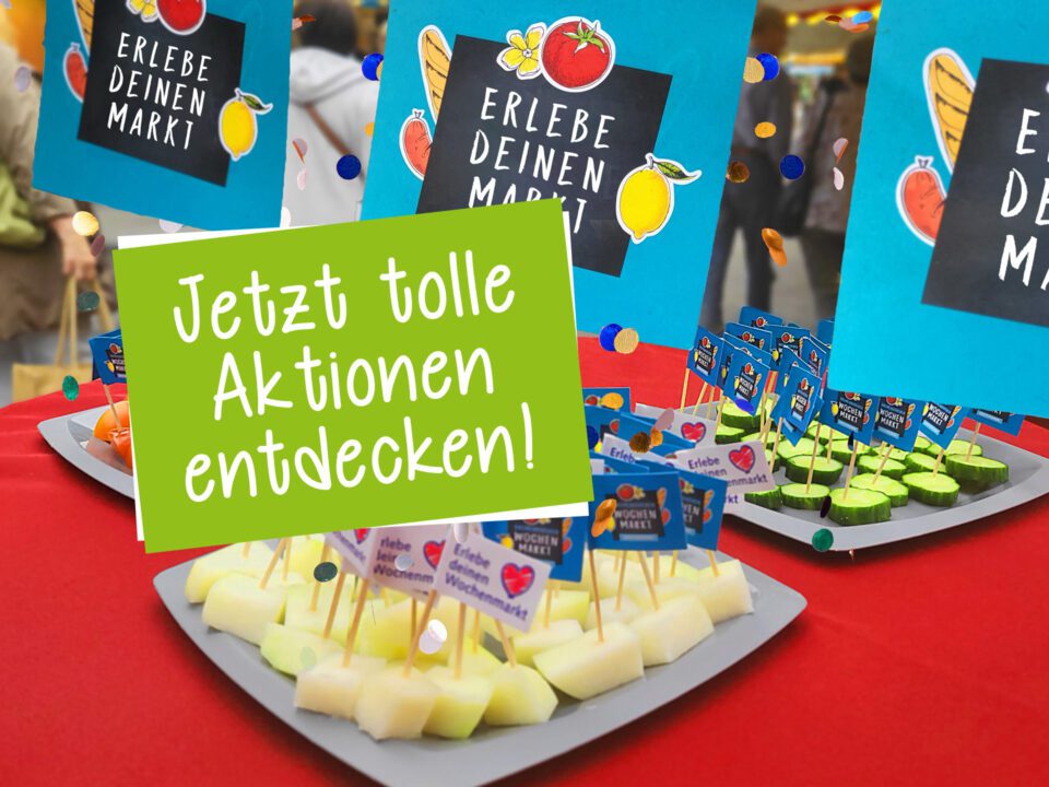 Eine Auslage mit Obst- und Gemüseproben auf Tabletts mit kleinen Schildern, die auf einem roten Tisch auf einem Markt aufgestellt sind. Auf den Schildern steht "Erlebe deinen Markt" und "Jetzt tolle Aktionen entdecken!" auf Deutsch. Im unscharfen Hintergrund sind Menschen zu sehen.