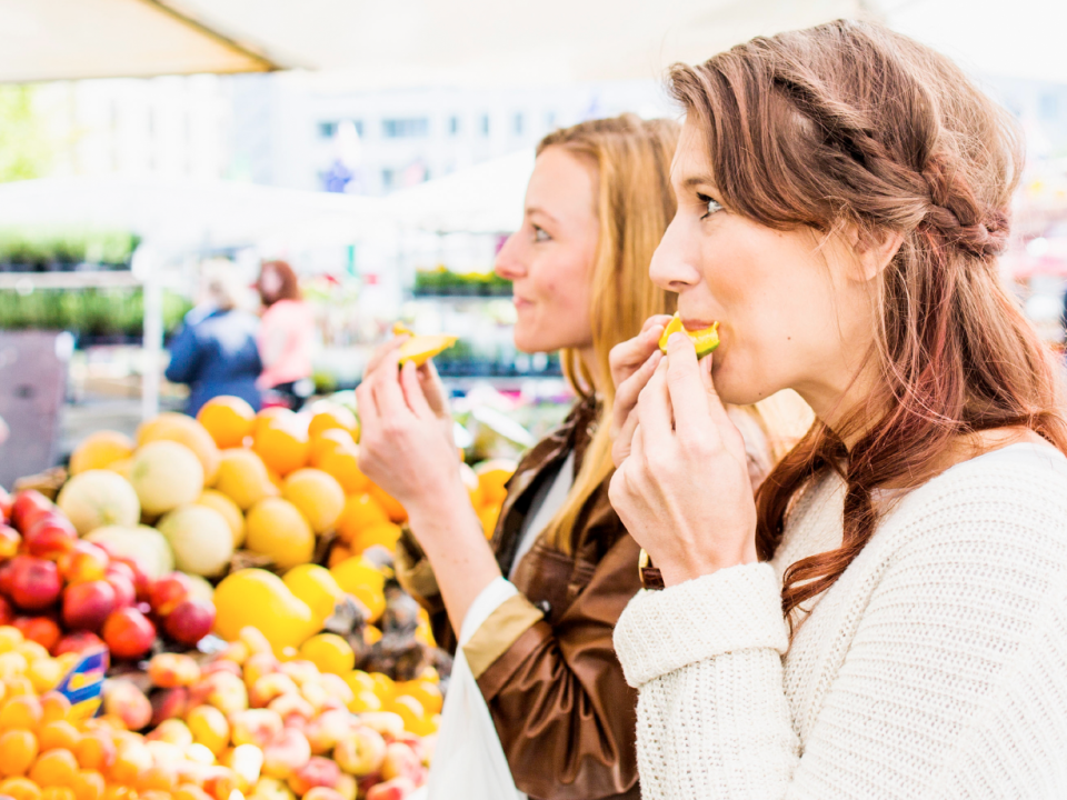 Zwei Frauen stehen an einem Marktstand im Freien und probieren frisches Obst. Im Vordergrund stehen farbenfrohe Produkte, darunter Pfirsiche und Melonen.